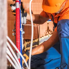 Instalator w pomarańczowym kasku i niebieskich spodniach wykonuje próbę ciśnieniową instalacji wodnej zgodnie z normą, pracując przy czerwonych i niebieskich rurach.