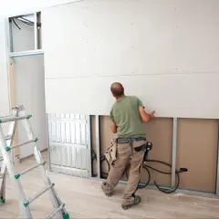 Pracownik wykonuje montaż płyt gipsowych na wkręty do stelaża. Obok stoi drabina.