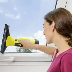 Kobieta z uśmiechem czyści okno żółtą myjką ciśnieniową do okien, usuwając smugi.