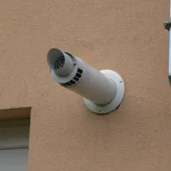 Biały komin gazowy przez ścianę budynku, obok metalowy kanał wentylacyjny.