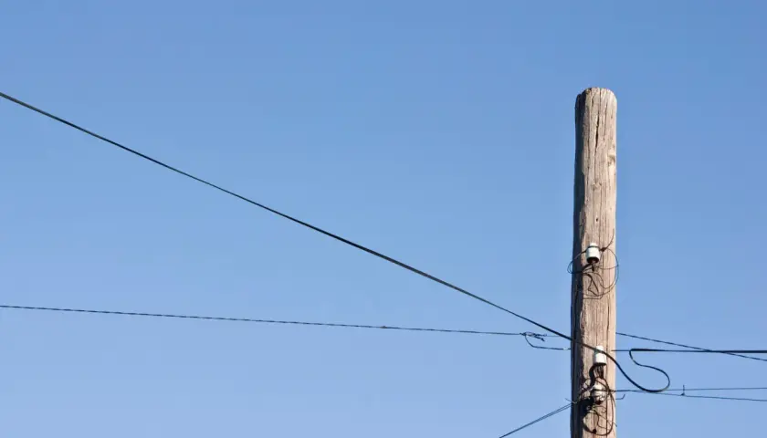 Drewniany słup z izolatorami, na którym widać światłowód na słupach. Linie energetyczne przecinają błękitne niebo.