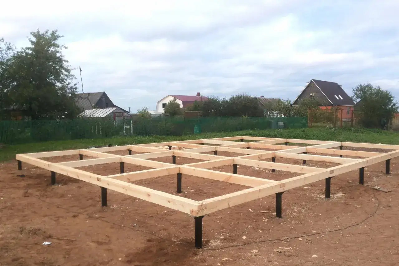 Drewniana konstrukcja na słupach, przygotowywana jako fundament pod dom. W tle widać domy i drzewa.
