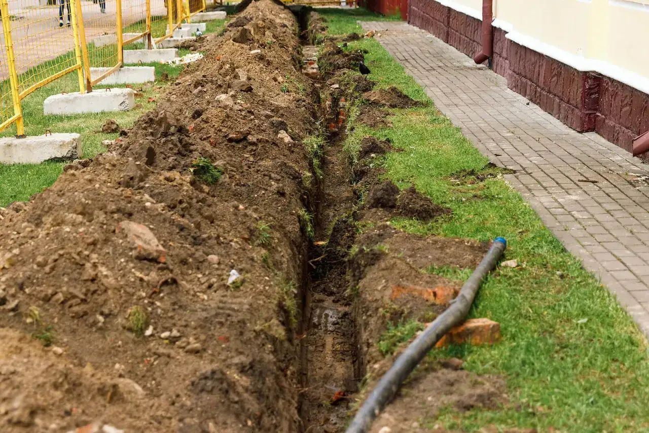 Trwa zabudowa rowu melioracyjnego. Wykop obok chodnika i budynku, z luźną ziemią i rurą.